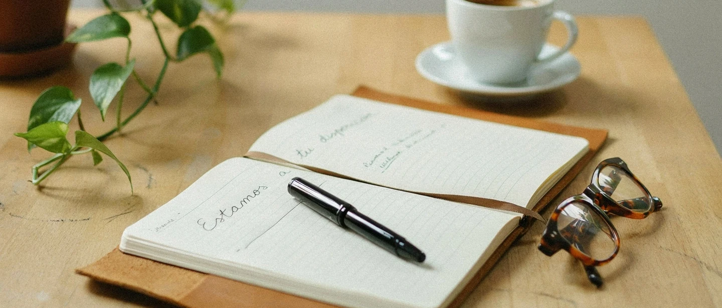 Cuaderno abierto con un bolígrafo, una taza de café y unas gafas sobre una mesa